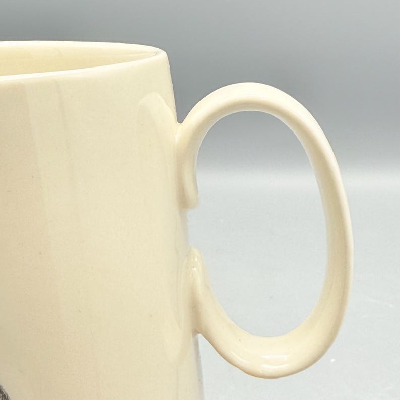 May include: Cream-colored ceramic mug with a curved handle. The mug has a smooth, glossy finish and a simple, classic design. The handle is large and easy to grip, making it suitable for hot or cold beverages.