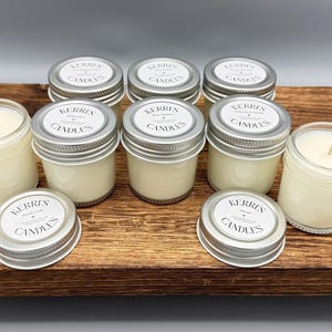 May include: A collection of small glass jar candles with silver metal lids. Each lid features a white circular label with the text Kerri's Candles. Some jars are open, revealing off-white wax and a centered wick, while others are sealed.