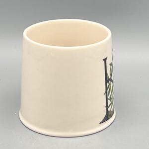 May include: Cream-colored ceramic mug with a decorative design. The design features a black letter "H" with green and white botanical accents. The mug has a slightly flared shape and a smooth, glossy finish.