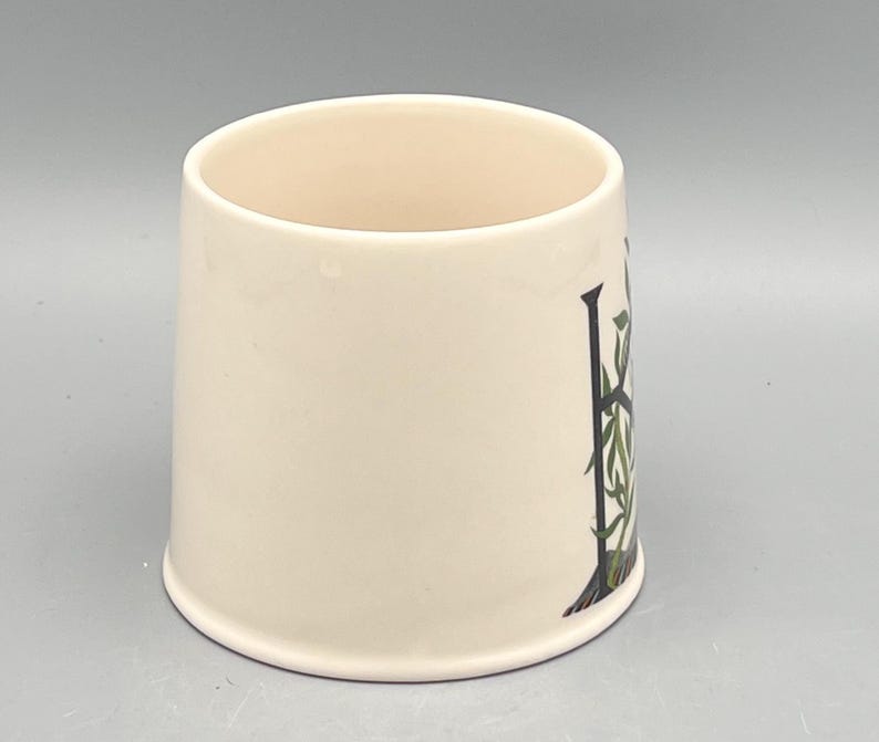 May include: Cream-colored ceramic mug with a decorative design. The design features a black letter "H" with green and white botanical accents. The mug has a slightly flared shape and a smooth, glossy finish.