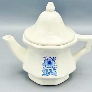 May include: Cream-colored ceramic teapot with a lid and handle. The teapot features a blue floral design on the front. The spout and handle are integrated into the design. The lid has a small knob on top.