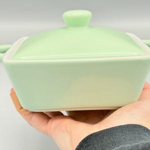 Puede incluir: Una fuente de cer&aacute;mica verde claro con tapa a juego. La fuente cuadrada tiene un borde blanco y asas peque&ntilde;as. La tapa tiene un asa redondeada. La fuente se sostiene en una mano.