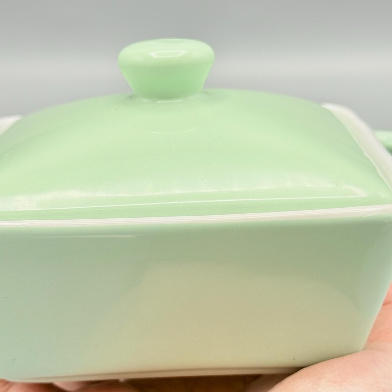 Puede incluir: Una fuente de cer&aacute;mica verde claro con tapa a juego. La fuente es cuadrada con esquinas redondeadas y un borde blanco. La tapa tiene un peque&ntilde;o asa redondeada. La fuente est&aacute; sostenida en la mano de una persona.