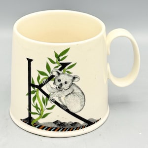 May include: Cream-colored ceramic mug featuring a black letter "K" with a koala bear illustration. The koala is perched on the letter with green leafy vines. The mug has a curved handle and a decorative band at the base.