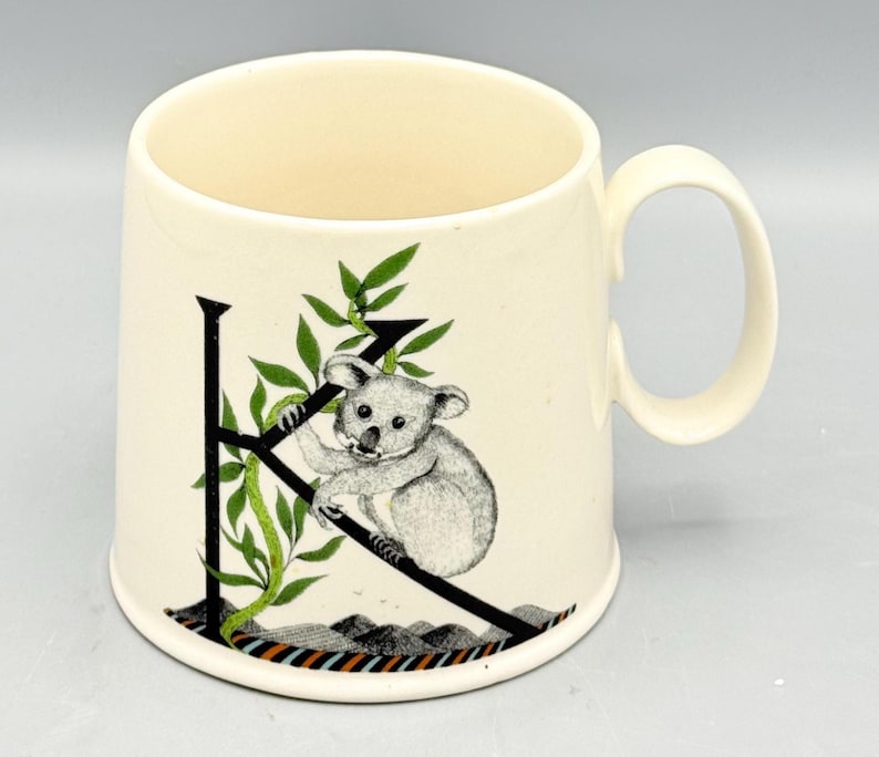 May include: Cream-colored ceramic mug featuring a black letter "K" with a koala bear illustration. The koala is perched on the letter with green leafy vines. The mug has a curved handle and a decorative band at the base.