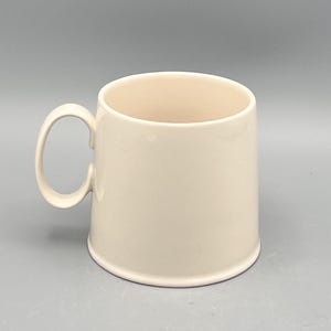 May include: A cream-colored ceramic mug with a rounded handle. The mug has a cylindrical shape and a smooth, glossy finish. The interior of the mug is a slightly lighter shade of cream. The mug is designed for hot or cold beverages.
