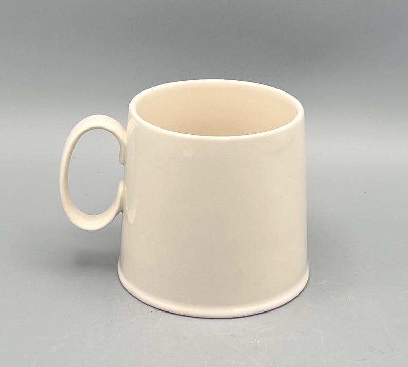 May include: A cream-colored ceramic mug with a rounded handle. The mug has a cylindrical shape and a smooth, glossy finish. The interior of the mug is a slightly lighter shade of cream. The mug is designed for hot or cold beverages.