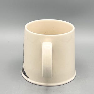 May include: Cream-colored ceramic mug with a white handle. The mug has a cylindrical shape and a simple design, suitable for coffee or tea. The mug is clean and appears unused.