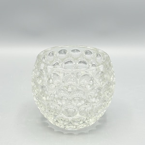 May include: A clear glass votive candle holder with a textured, bubble-like pattern. The round, open-top container is designed to hold a small candle. The glass is transparent, allowing light to pass through.
