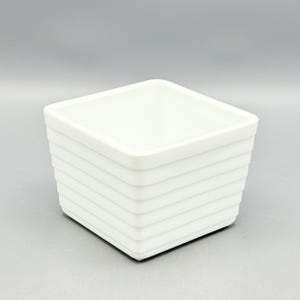 May include: A square white ceramic bowl featuring a series of horizontal ridges along its exterior walls. The vessel has a clean, open top design with a smooth finish, showcasing a uniform white color throughout its entire structure.
