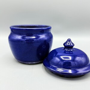 May include: A deep cobalt blue ceramic jar with a matching removable lid. The jar features a rounded, bulbous body with a smooth, glossy finish. The lid has a small, pointed finial handle at the center, reflecting light across its curved surface.