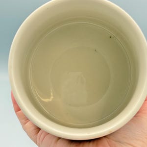May include: A cream-colored ceramic bowl, viewed from above, with a smooth interior and a slightly rounded rim. The bowl's interior has a subtle, light green hue. The bowl is held by a hand, with a light blue background.