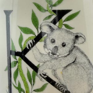 May include: A detailed illustration of a koala bear clinging to a black branch, surrounded by green leaves. The koala is gray with a black nose and large, round eyes. The letter "J" is visible in the background.