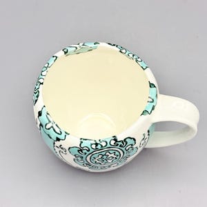 May include: A white ceramic mug with a teal and black floral design. The mug has a rounded shape and a handle.