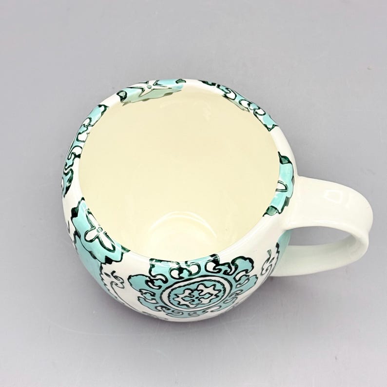May include: A white ceramic mug with a teal and black floral design. The mug has a rounded shape and a handle.