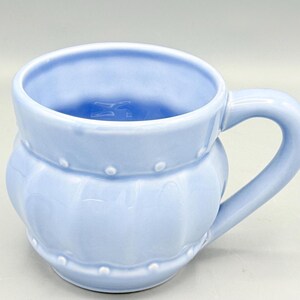 May include: A light blue ceramic mug with a rounded body and a curved handle. The mug features a decorative design with small, raised dots around the middle and top. The interior of the mug is also light blue.