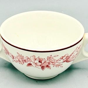 May include: A cream-colored ceramic soup bowl featuring two side handles and a delicate maroon floral pattern encircling the exterior. The rim is accented with a thin maroon line, and the interior of the bowl is plain, smooth, and off-white.