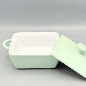 Puede incluir: Un plato cuadrado de cer&aacute;mica verde claro con tapa a juego. El plato tiene un interior blanco y peque&ntilde;as asas a cada lado. La tapa est&aacute; ligeramente entreabierta, revelando el interior del plato. El plato es probablemente para servir o almacenar alimentos.