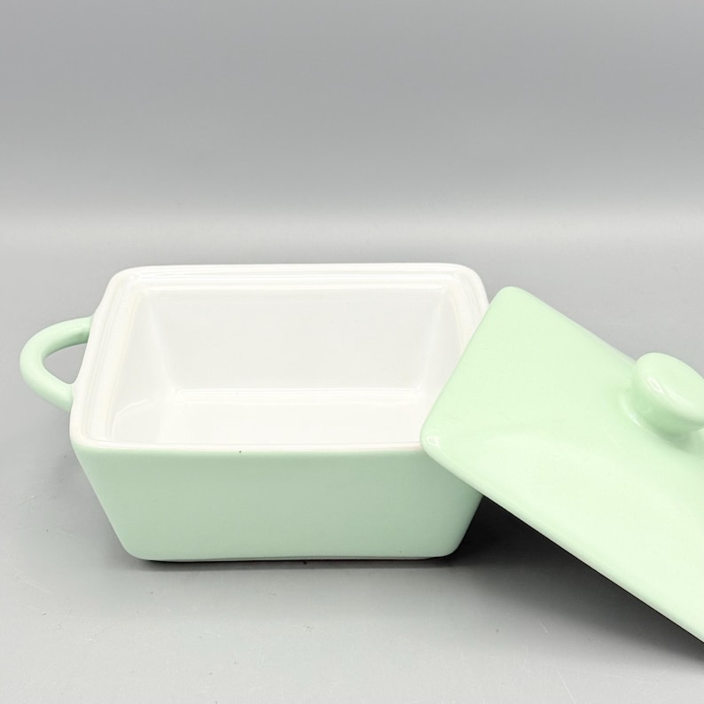 Puede incluir: Un plato cuadrado de cer&aacute;mica verde claro con tapa a juego. El plato tiene un interior blanco y peque&ntilde;as asas a cada lado. La tapa est&aacute; ligeramente entreabierta, revelando el interior del plato. El plato es probablemente para servir o almacenar alimentos.