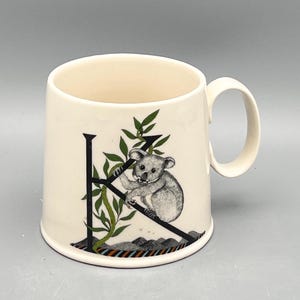 May include: Cream-colored ceramic mug featuring a black letter "K" with a koala bear illustration. The koala is perched on a branch with green leaves. The mug has a curved handle and a colorful striped design at the base.