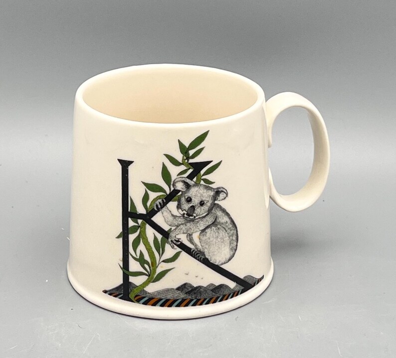May include: Cream-colored ceramic mug featuring a black letter "K" with a koala bear illustration. The koala is perched on a branch with green leaves. The mug has a curved handle and a colorful striped design at the base.