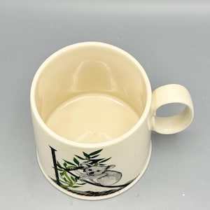 May include: Cream-colored ceramic mug with a rounded handle. The mug features a black and green illustration of a koala bear on a branch. The interior of the mug is a matching cream color.