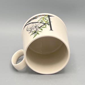 May include: Cream-colored ceramic mug featuring a black letter "A" with a koala bear illustration. The koala is hanging from a branch with green leaves. The mug has a curved handle and a smooth, glossy finish.