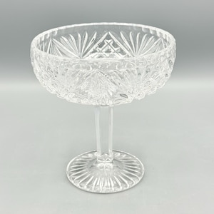 May include: A clear, stemmed glass champagne coupe with a wide, shallow bowl and a decorative, cut-glass pattern. The bowl sits atop a slender stem and a circular base. The glass is designed with a starburst pattern on the base and a detailed design around the bowl.