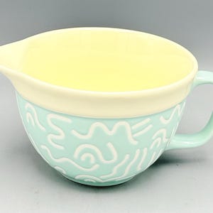 May include: A ceramic mixing bowl with a handle and a pouring spout. The exterior is light mint green with a white squiggly line pattern, while the interior and the upper rim are a solid pale yellow. The bowl features a rounded base and side.