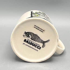 May include: White ceramic mug with a black and white fish illustration on the bottom. The mug has a handle and a design of bamboo and leaves. The text "BALDUCCI for ANTHROPOLOGIE" is printed on the bottom.