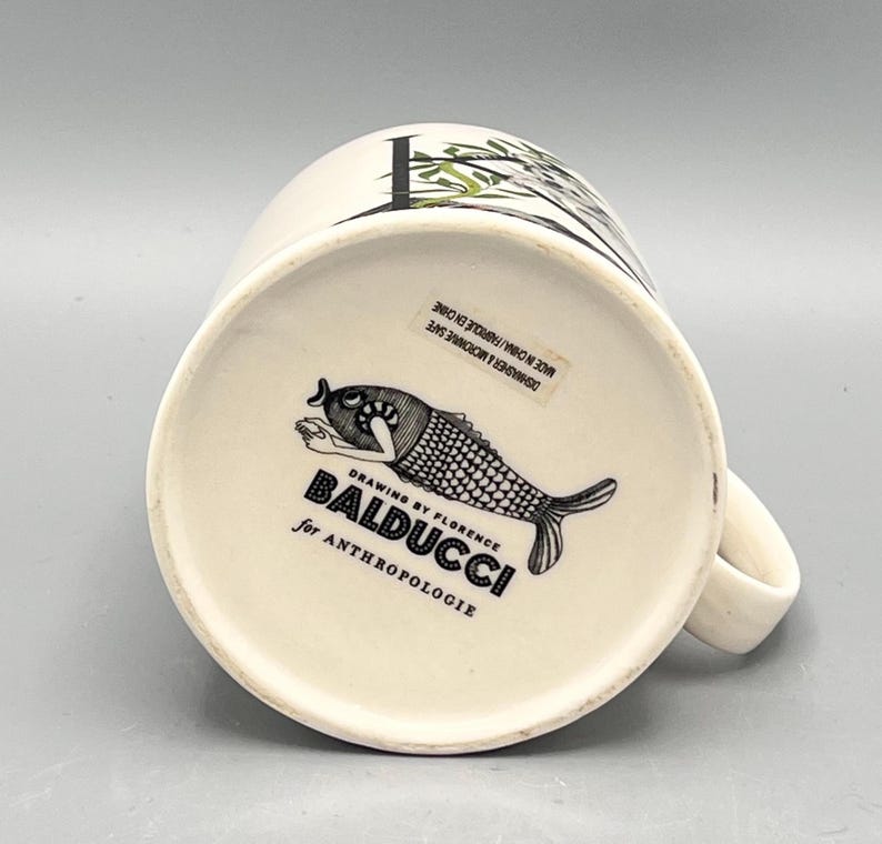May include: White ceramic mug with a black and white fish illustration on the bottom. The mug has a handle and a design of bamboo and leaves. The text "BALDUCCI for ANTHROPOLOGIE" is printed on the bottom.