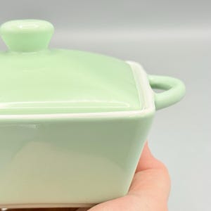Puede incluir: Una fuente de cer&aacute;mica verde claro con tapa y asa. La fuente es cuadrada con esquinas redondeadas y un borde blanco. La tapa tiene un asa peque&ntilde;a y redonda. La fuente se sostiene en una mano.