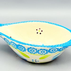 May include: A ceramic bowl shaped like a fish with a cream-colored interior featuring small purple and yellow floral accents. The exterior rim is bright blue with a textured pattern, while the body displays blue floral motifs and green leaves.