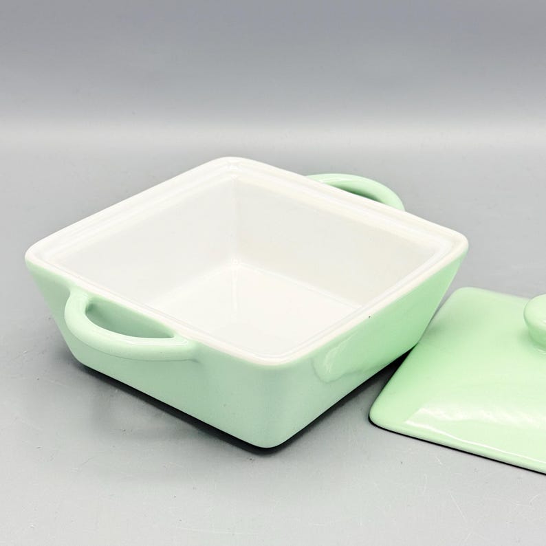Puede incluir: Una fuente para hornear cuadrada de cer&aacute;mica verde claro con interior blanco. La fuente tiene dos asas curvas y una tapa a juego con un peque&ntilde;o pomo. La tapa est&aacute; parcialmente quitada.