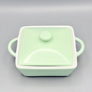 Puede incluir: Un plato cuadrado de cer&aacute;mica verde claro con tapa a juego y dos asas curvas. La tapa tiene un peque&ntilde;o pomo redondo. El plato tiene un borde blanco y se asienta sobre una superficie gris claro. El plato est&aacute; dise&ntilde;ado para servir.
