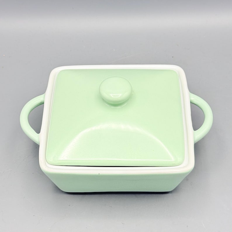 Puede incluir: Un plato cuadrado de cer&aacute;mica verde claro con tapa a juego y dos asas curvas. La tapa tiene un peque&ntilde;o pomo redondo. El plato tiene un borde blanco y se asienta sobre una superficie gris claro. El plato est&aacute; dise&ntilde;ado para servir.