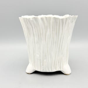 May include: A white ceramic planter with a textured, vertical-lined design. The planter has a flared top and sits on three small, rounded feet. The overall shape is a trapezoid. The planter is suitable for indoor or outdoor use.