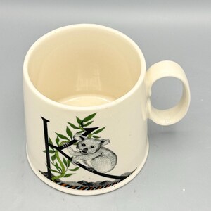 May include: Cream-colored ceramic mug with a rounded handle. The mug features a black letter "K" with a koala bear illustration and green leaves. The koala is gray and white. The mug has a smooth, glossy finish.