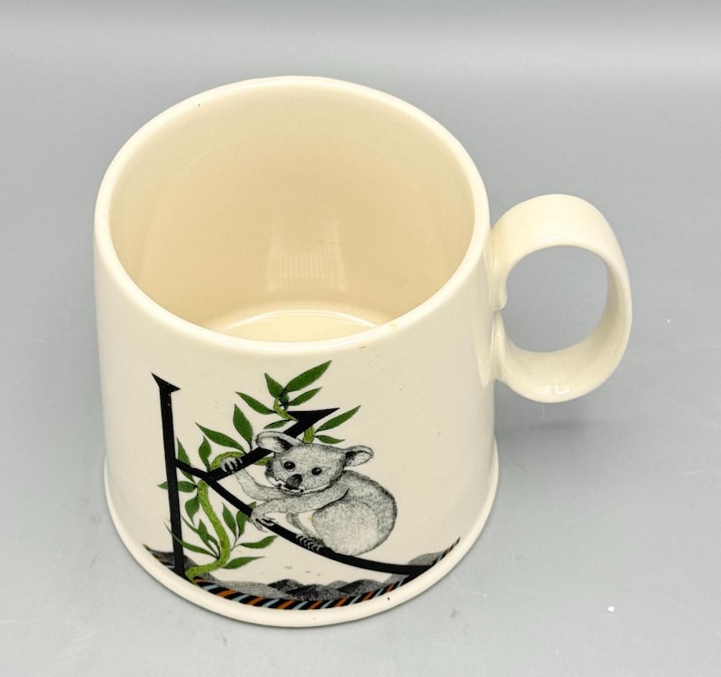 May include: Cream-colored ceramic mug with a rounded handle. The mug features a black letter "K" with a koala bear illustration and green leaves. The koala is gray and white. The mug has a smooth, glossy finish.