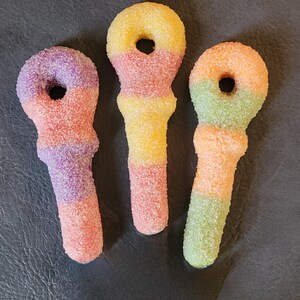 Può includere: Tre caramelle gommose colorate e ricoperte di zucchero a forma di microfono. Ogni caramella ha una combinazione di colori diversa: viola e rosa, giallo e rosa e arancione e verde. Le caramelle sono su una superficie scura.