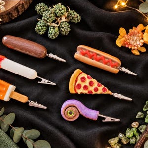 May include: A collection of six food-themed memo clips featuring a brown sausage, a white stick with a red tip, an orange popsicle, a hot dog with red ketchup, a pepperoni pizza slice, and a purple spiral party blower, each with a metal clip.