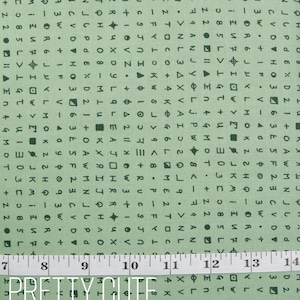 May include: Green fabric with a repeating pattern of letters, numbers, and symbols. The fabric has a textured surface and a ruler is visible at the bottom of the image. The text "PRETTY CUTE QUILTS" is printed in black on a white background.