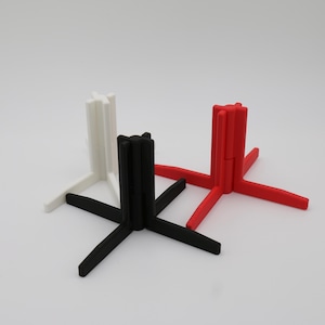 Op de afbeelding: Drie 3D-geprinte standaards in wit, zwart en rood. Elke standaard heeft een vierkante basis en vier armen die zich uitstrekken vanaf de basis. De armen zijn verbonden met een centrale kolom.