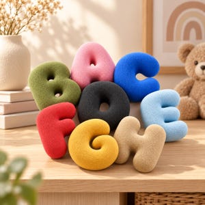Op de afbeelding: Een verzameling van zachte pluche alfabetletters, waaronder A, B, C, E, F, G en H, in diverse kleuren zoals olijfgroen, roze, blauw, zwart, rood, geel en beige, gerangschikt op een houten ondergrond.