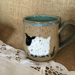 May include: A brown ceramic mug with a blue rim and handle. The mug has a white sheep with black face and legs painted on the front.