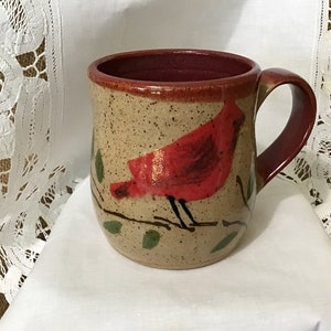May include: Handmade ceramic mug with a red cardinal bird design. The mug features a speckled beige exterior, a burgundy interior, and a matching handle. The bird is perched on a branch with green leaves. A unique piece for coffee or tea.