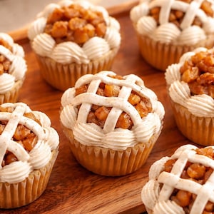 Puede incluir: Una colección de cupcakes de tarta de manzana con un relleno de manzana dorada en dados, cubiertos con un enrejado de glaseado beige claro y un borde de crema, todo dentro de moldes de papel plisado para hornear.