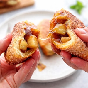 Puede incluir: Un pastel frito de color dorado partido por la mitad, que revela un relleno de manzana cálido y pegajoso con un espeso glaseado de caramelo. El exterior está cubierto por una capa gruesa de azúcar y canela, con un hilo de almíbar estirado entre ambas partes.