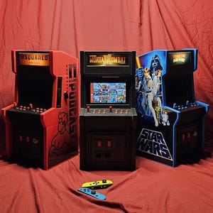Pode incluir: Três máquinas de arcade em miniatura. Uma é vermelha com o texto "DSQUARED PODCAST", outra é preta com o texto "MORTAL KOMBAT", e a terceira é azul com o texto "STAR WARS". Dois controles de jogo estão em primeiro plano.