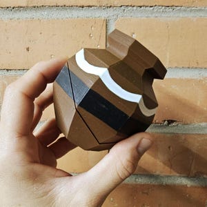 May include: A geometric, brown, 3D-printed object with black and white accents. The object is held in a hand and has a unique, angular design. The background is a brick wall.
